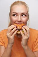 positivo Affamato giovane bello bionda signora con casuale acconciatura guardare allegramente a parte mentre mangiare Hamburger con grande piacere, in piedi al di sopra di bianca sfondo nel arancia maglietta foto