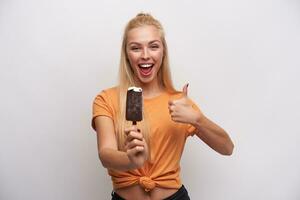 contento attraente giovane femmina con lungo bionda capelli conservazione ghiaccio crema nel sollevato mano e mostrando pollice mentre in piedi al di sopra di bianca sfondo, essere nel alto spirito e sorridente a grandi linee foto