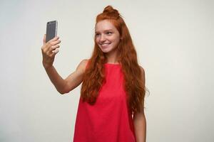 studio foto di positivo giovane lettore donna con ondulato rosso capelli in posa al di sopra di bianca sfondo senza rendere su, fabbricazione autoscatto su sua smartphone nel casuale rosa vestito