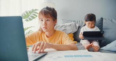 felice allegra famiglia asia mamma che usa il laptop con il suo bambino tiene il fumetto di interesse per tablet sul divano mentre lavora da casa in soggiorno. distanziamento sociale, quarantena per la prevenzione del coronavirus. foto