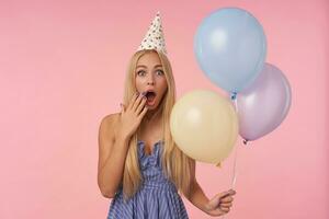 studio tiro di giovane lungo dai capelli donna nel festivo Abiti e cono cappello Tenere mazzo di elio palloncini mentre in posa al di sopra di rosa sfondo, guardare a telecamera sorprendentemente e copertura bocca con palma foto