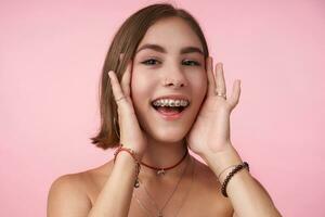 allegro giovane bello dagli occhi marroni brunetta donna con corto taglio di capelli Tenere sua viso con sollevato mani e sorridente felicemente mentre in posa al di sopra di rosa sfondo foto