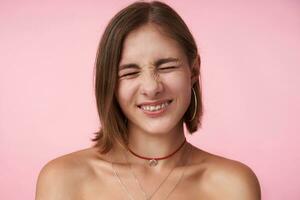 ritratto di piacevole guardare giovane corto dai capelli signora con naturale trucco accigliato sua viso mentre sorridente allegramente con chiuso occhi, isolato al di sopra di rosa sfondo foto