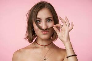 positivo giovane corto dai capelli brunetta donna con naturale trucco imitando baffi con sua capelli mentre fabbricazione divertimento, essere nel simpatico umore mentre in piedi al di sopra di rosa sfondo foto