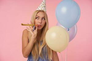 bellissimo bionda lungo dai capelli signora conservazione festa corno nel sua bocca e guardare a parte positivamente, in posa nel multicolore aria palloncini mentre festeggiare compleanno, in piedi al di sopra di rosa sfondo foto