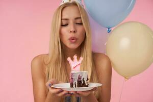 positivo bella giovane lungo dai capelli femmina festeggiare compleanno con multicolore aria palloncini, conservazione compleanno torta nel mani e soffiaggio su candela, in piedi al di sopra di rosa sfondo foto