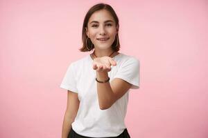 piacevole guardare giovane positivo dagli occhi marroni brunetta femmina sorridente leggermente mentre raccolta sua palma, indossare di base bianca maglietta mentre in posa al di sopra di rosa sfondo foto