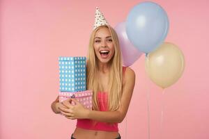 gioioso attraente giovane lungo dai capelli donna nel rosa superiore e vacanza berretto essere eccitato e sorpreso per ottenere compleanno regali, in posa al di sopra di rosa sfondo nel multicolore aria palloncini foto