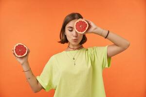 femmina alunno, giovane signora con corto brunetta capelli, sognare con frutta. Tenere pompelmo al di sopra di sua occhio, copertina uno occhio. in piedi al di sopra di arancia sfondo. indossare verde maglietta, bretelle e bracciali foto