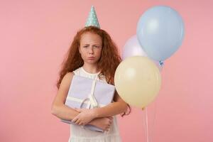 studio tiro di irritato testa Rossa femmina ragazzo con lungo Riccio capelli accigliato per telecamera e pieghevole labbra, in posa al di sopra di rosa sfondo nel festivo Abiti e compleanno cap. bambini e celebrazione concetto foto