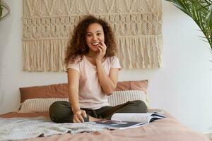 giovane africano americano sorpreso donna con Riccio capelli, si siede su il letto e tocchi guancia, a grandi linee sorrisi e legge un' nuovo rivista, godere freddo notizia nel rivista, freddo spende gratuito tempo a casa. foto