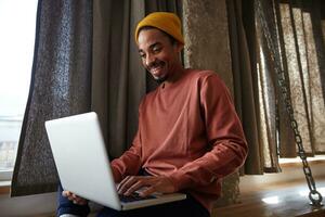 ritratto di gioioso bello buio spellato uomo seduta di finestra e conservazione il computer portatile su il suo gambe, guardare felicemente su schermo e sorridente ampiamente, vestito nel casuale Abiti foto