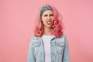 oltraggiato ragazza con rosa capelli Spettacoli sua dispiacere di scoprendo sua denti, sembra scontento a il telecamera, sta al di sopra di rosa sfondo. foto