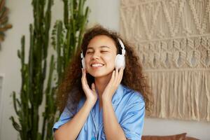 contento Riccio mulatto giovane signora ubicazione su il letto, a grandi linee sorrisi e chiuso occhi, ascoltando preferito canzone nel cuffia, godendo il musica e domeniche mattina. foto