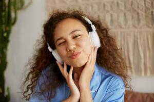 contento Riccio mulatto giovane signora ubicazione su il letto, occhiolino e ascoltando preferito musica nel cuffia, godendo il domeniche mattina. foto