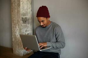 interno tiro di concentrato bello buio spellato tipo con barba, Tenere il computer portatile e guardare su schermo con riflessivo viso, seduta al di sopra di bianca parete nel casuale Abiti foto