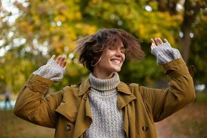 lieta bene guardare giovane Marrone dai capelli femmina agitando sua corto capelli e conservazione mani sollevato, sorridente a grandi linee con chiuso occhi mentre in piedi al di sopra di parco sfondo foto