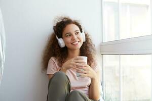 allegro giovane africano americano donna con Riccio capelli seduta di il finestra, ascoltando preferito musica a cuffie e potabile Tè, sembra a il finestra e godendo giorno a casa. foto