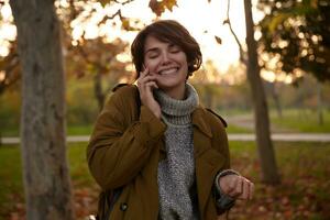 piacevole guardare giovane bello Marrone dai capelli donna con peso acconciatura conservazione sua occhi chiuso mentre sorridente Cordiali saluti, avendo simpatico Telefono conversione mentre a piedi al di sopra di città giardino foto