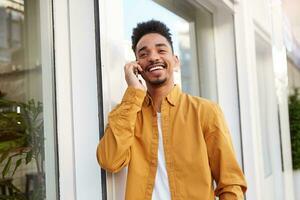 ritratto di giovane africano americano positivo tipo, a piedi giù il strada e parlando su il Telefono con il suo amico a grandi linee sorridente e sembra lontano, godendo il giorno. foto