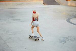 giovane tatuato signora con bionda capelli raid su skateboard, indossa nel un' rosso maglietta e denim corti, con un' a maglia bandana su sua testa, godendo il giorno nel pattinare parco. foto