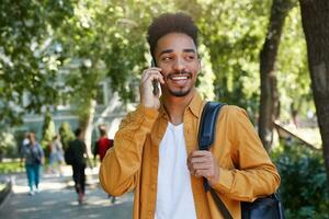 ritratto di giovane buio spellato sorridente tipo indossa nel un' giallo camicia e un' bianca maglietta con un' zaino su uno spalla, a piedi nel il parco e parlando su il Telefono, sorridente e gode il giorno. foto