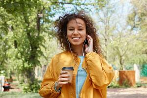 ahah carino Riccio buio spellato signora a grandi linee sorridente, indossare un' giallo giacca, a piedi nel il parco, Tenere un' tazza di caffè, ascoltare per musica, e godendo il tempo atmosferico. foto