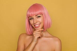 ritratto di giovane contento occhi azzurri rosa dai capelli femmina guardare felicemente a telecamera con affascinante Sorridi, Tenere sollevato mano su sua mento mentre in posa al di sopra di mostarda sfondo foto