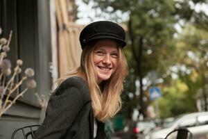 ritratto di giovane piacevole guardare femmina con lungo bionda capelli in posa al di sopra di città sfondo su soleggiato fine settimana, essere nel simpatico umore e sorridente Cordiali saluti foto