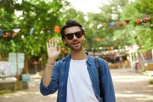 attraente giovane buio dai capelli uomo a piedi attraverso città giardino e raccolta mano nel benvenuto gesto, indossare casuale Abiti e occhiali da sole foto