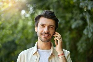 all'aperto avvicinamento di affascinante giovane buio dai capelli uomo con barba avendo parlare su il suo mobile Telefono mentre a piedi lungo verde giardino su soleggiato giorno foto