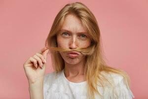 avvicinamento di giovane bella testa Rossa femmina con casuale acconciatura imitando baffi con serratura di capelli e guardare sul serio a telecamera, vestito nel casuale Abiti mentre in posa al di sopra di rosa sfondo foto