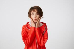 foto di giovane carino corto dai capelli ragazza vestito nel bianca golf e rosso pioggia cappotto, guardare a il telecamera con stupito espressione, Spedire bacio, palme su guance, in piedi al di sopra di bianca sfondo.