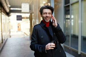 gioioso giovane attraente Marrone dai capelli maschio con di moda taglio di capelli ridendo felicemente mentre avendo Telefono conversazione con amico, isolato al di sopra di urbano sfondo foto