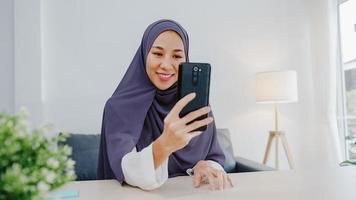 giovane donna d'affari musulmana asiatica che utilizza lo smartphone parla con un amico tramite videochat brainstorming riunione online mentre lavora a distanza da casa in soggiorno. distanziamento sociale, quarantena per il virus corona. foto