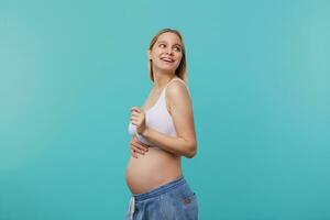 lato Visualizza di giovane attraente dalla testa bianca futuro mamma guardare felicemente al di sopra di sua spalla e Tenere sollevato mano su sua gonfiarsi, isolato al di sopra di blu sfondo foto