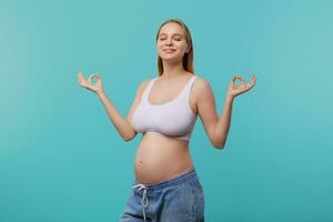 bene guardare giovane positivo dalla testa bianca incinta donna con naturale trucco sorridente piacevolmente con chiuso occhi e raccolta mani con mudra gesto, isolato al di sopra di blu sfondo foto