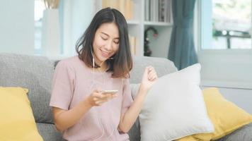 donna asiatica che ascolta musica e usa lo smartphone, donna che usa il tempo di relax sdraiata sul divano di casa nel soggiorno di casa. musica d'ascolto femminile felice con il concetto delle cuffie.