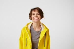 foto di contento giovane simpatico donna nel giallo pioggia cappotto, godere il vita, sembra a il telecamera con positivo espressioni, a grandi linee sorridente al di sopra di bianca parete.