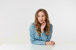 foto di giovane bellissimo donna con lungo biondo ondulato capelli, indossare un' denim camicia, ubicazione a il tavolo oggetti di scena il suo mento con il suo mano, pensiero di qualcosa guardare lontano isolato al di sopra di bianca sfondo.