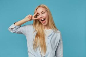 ritratto di contento giovane bionda femmina con lungo capelli indossare blu camicia e grigio maglietta, Tenere dita nel pace gesto mentre in piedi al di sopra di blu sfondo, sorridente a grandi linee a telecamera foto