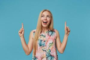 interno tiro di positivo bionda donna con lungo capelli puntamento verso l'alto con indice dita, in piedi al di sopra di blu sfondo nel fiorito vestire, sorridente felicemente e dimostrando sua bianca Perfetto denti foto