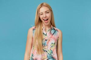 allegro attraente giovane bionda femmina con lungo capelli indossare fiorito camicia, in piedi al di sopra di blu sfondo con mani fuori uso, strizzando l'occhio a telecamera con largo gioioso Sorridi foto