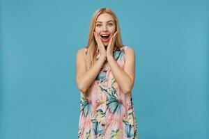 studio foto di sorpreso giovane lungo dai capelli bionda signora nel fiorito vestito in piedi al di sopra di blu sfondo, Tenere mani su sua guance e guardare a telecamera sorprendentemente