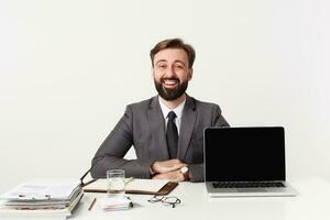 gioioso giovane bella barbuto uomo nel grigio completo da uomo e cravatta Lavorando a ufficio con moderno il computer portatile e taccuino, pieghevole mani su tavolo e sorridente allegramente mentre guardare per telecamera foto