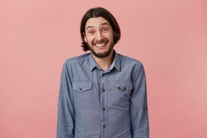 ritratto di allegro barbuto giovane uomo con lungo pettinato buio capelli nel un' bene umore, lieto, alza le spalle e sorridente isolato al di sopra di rosa sfondo. foto