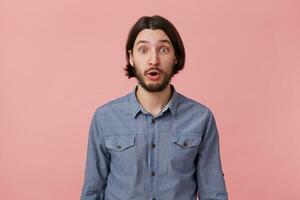 foto di stupito barbuto giovane uomo con lungo pettinato buio capelli nel denim camicia, isolato al di sopra di rosa sfondo.