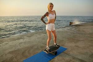 soleggiato foto di bella sportivo giovane donna con lungo bionda capelli pettinato nel treccia fabbricazione esercizi su equilibrio asse, in posa al di sopra di mare lungomare nel bianca superiore e pantaloncini