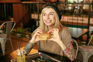 orizzontale tiro di bella tatuato bionda femmina nel beige maglietta e pelle nero berretto in posa al di sopra di moderno bar interno, guardare a parte con largo affascinante Sorridi e Tenere hamburger nel mani foto