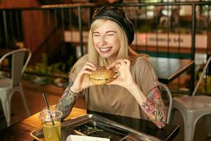 contento giovane bionda donna con tatuaggi avendo pranzo nel città bar, seduta a tavolo con hamburger nel mani e ridendo con gioia con chiuso occhi, indossare di moda Abiti foto